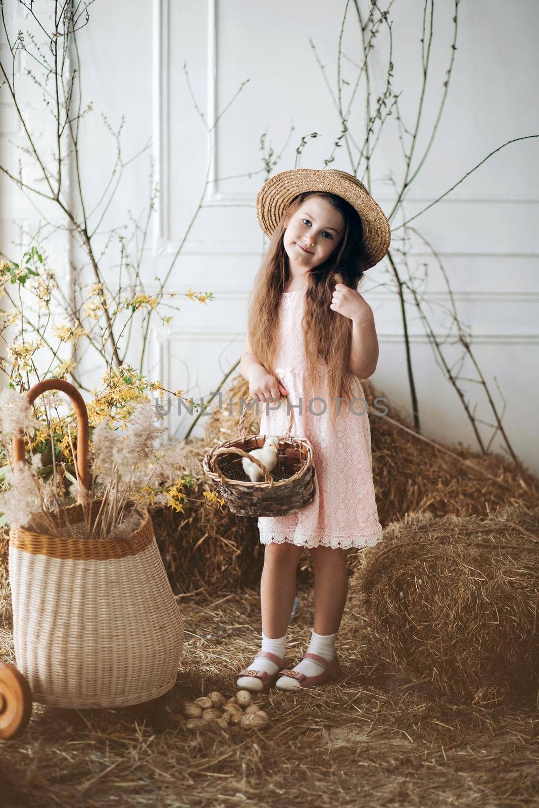 Front view of beautiful girl wearing pink dress and hay hat keeping basket with little chick. Cute female child looking aside and smiling while walking in garden in village. Concept of childhood.