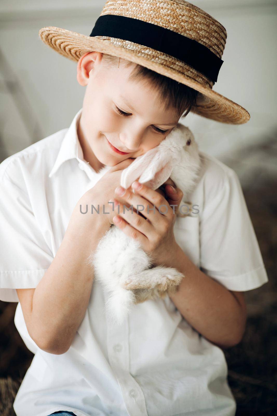 Boy in hay hat keeping cute white bunny and smiling by StudioLucky