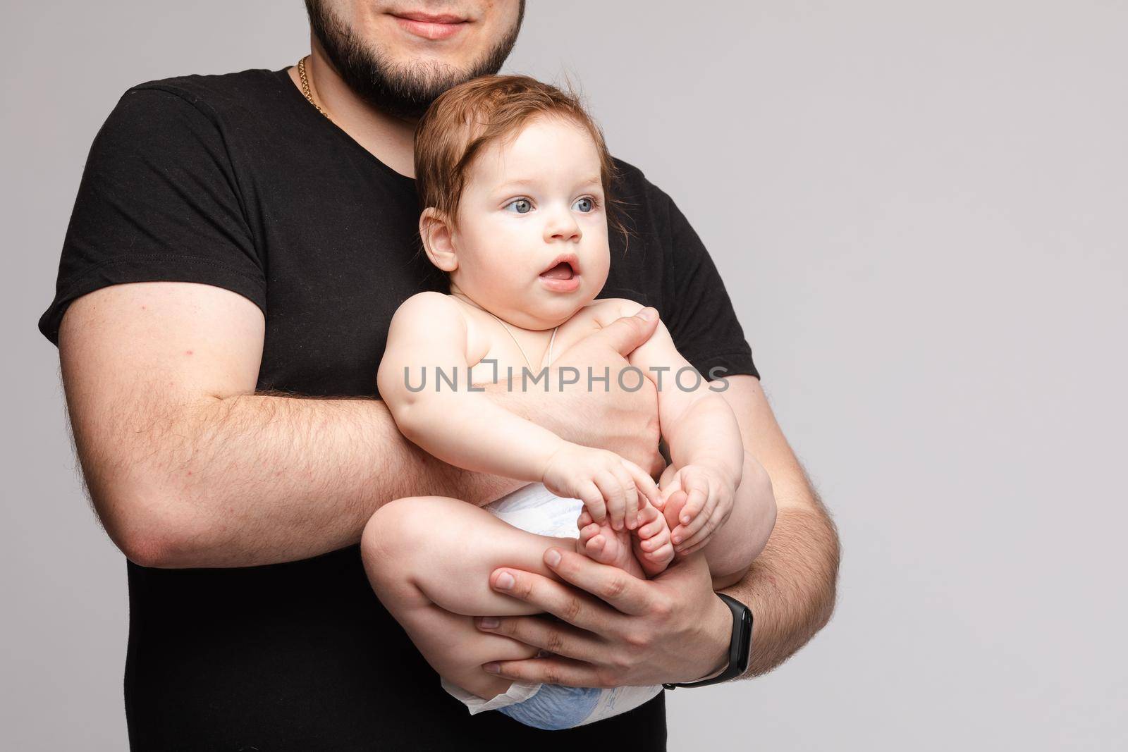 Side view of lovely father keeping little baby in hands and kissing kid on grey isolated background in studio. Happy male parent hugging and playing with child. Concept of family and love.