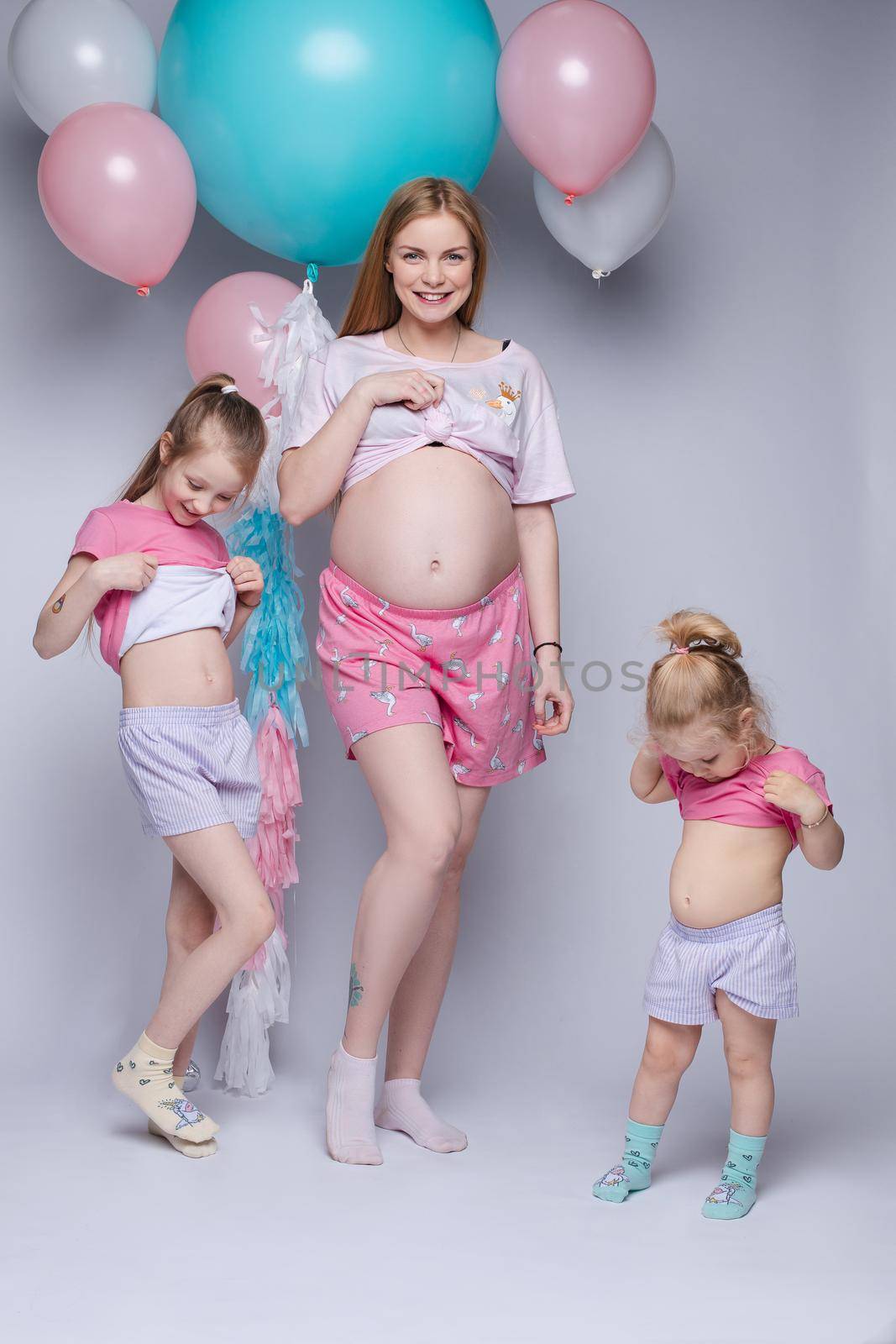 Stunning pregnant woman showing her belly. Her two daughters looking at their tummy too. They all wearing pyjamas. Studio portrait of mother and two daughters demonstrating bellies.