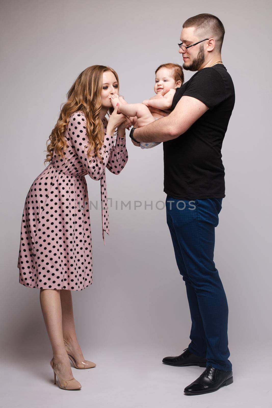 Portrait of happy family from three people wearing same coloured clothes. Blonde beautiful mother smiling and holding on hands surprised child. Handsome father in glasses hugging lovely wife and son.