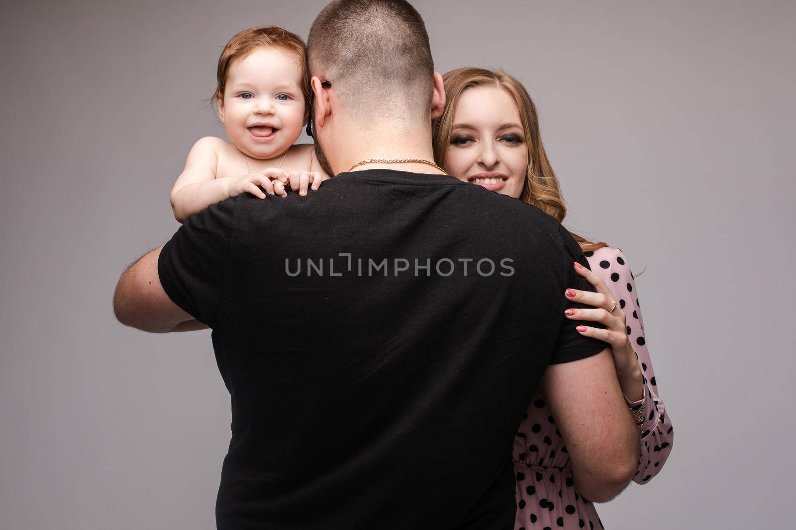 Back view stock photo of unrecognizable father with strong broad back embracing his beautiful smiling wife and holding their red-haired baby. Son and mother smiling at camera. Isolate on grey.