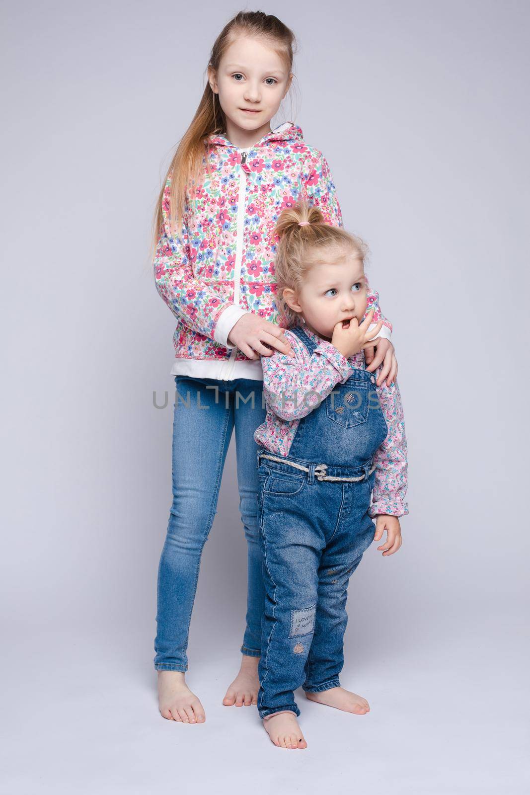 Front view of two pretty sisters keeping hands, looking at camera and smiling on isolated background in studio. Happy kids wearing stylish outfits posing together. Concept of friendship and fashion.