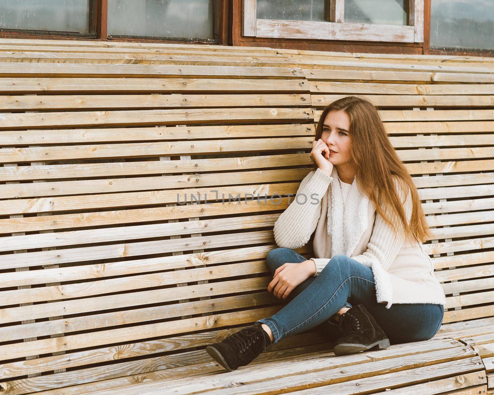 Beautiful young girl with long brown hair sits on a wooden bench made of planks and rests, relaxes and reflects. Outdoor photo shoot with attractive woman in winter or autumn