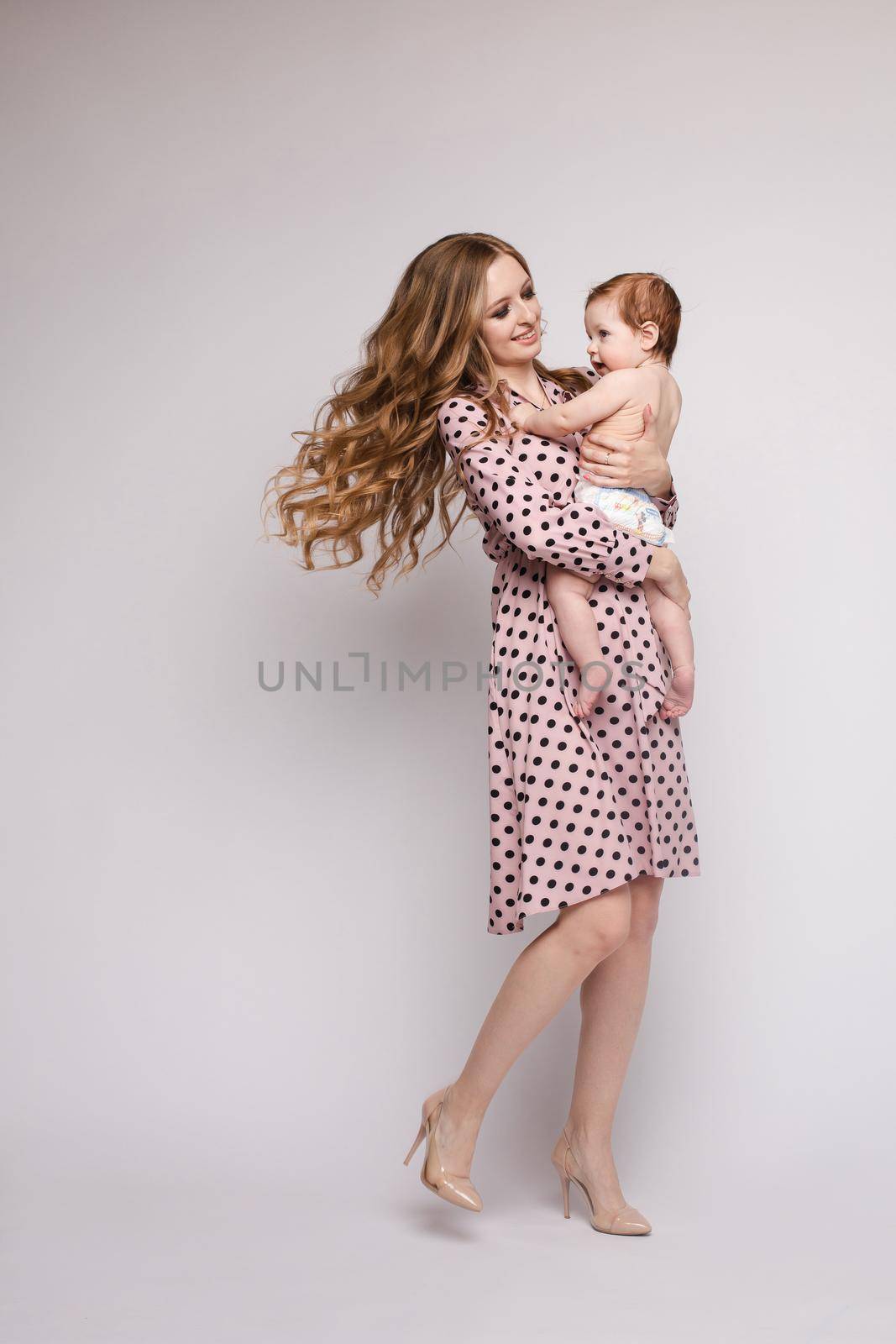 Portrait of lovely mother kissing little child on isolated background in studio. Pretty woman keeping cheerful daughter on hands and hugging her. Concept of family love and childhood.