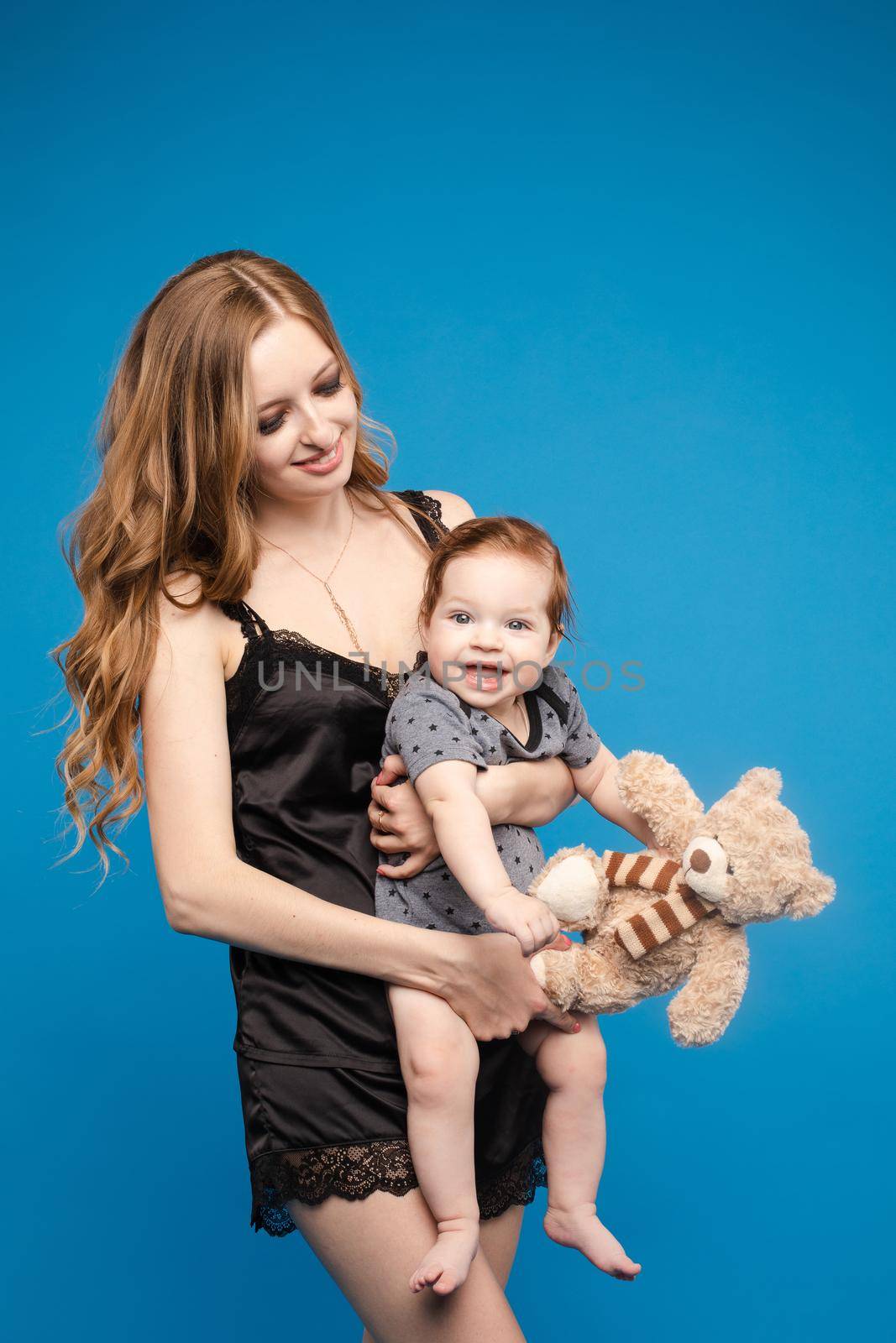 Front view of positive little baby laughing with mother in studio. Beautiful woman in black pajamas keeping cute daughter on hands. looking at camera and posing on blue isolated background.