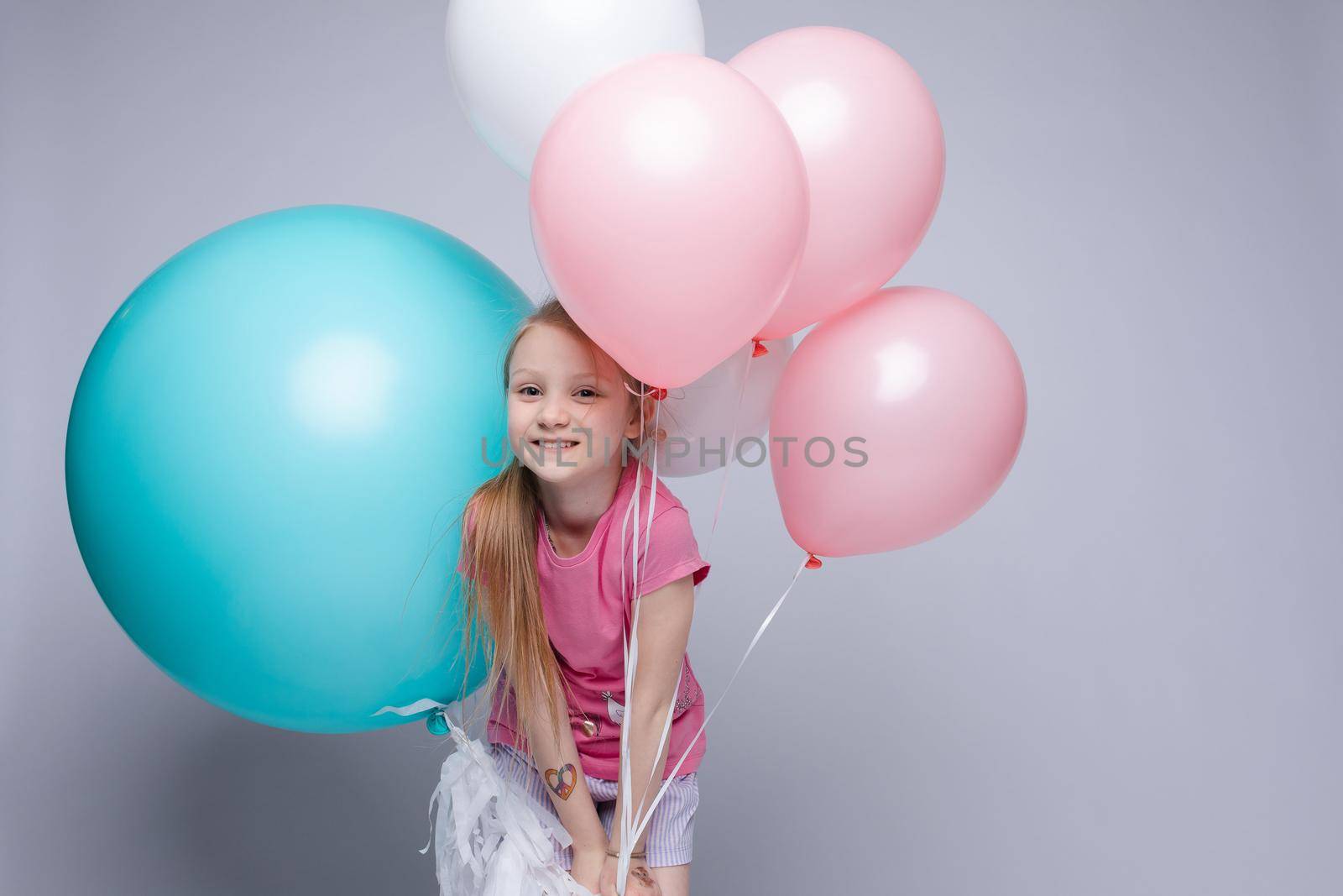 Studio portrait of lovely little girl with red or fair hair and freckles in pink t-shirt and shorts posing with toothy smile with pink, blue and white air balloons against grey background.