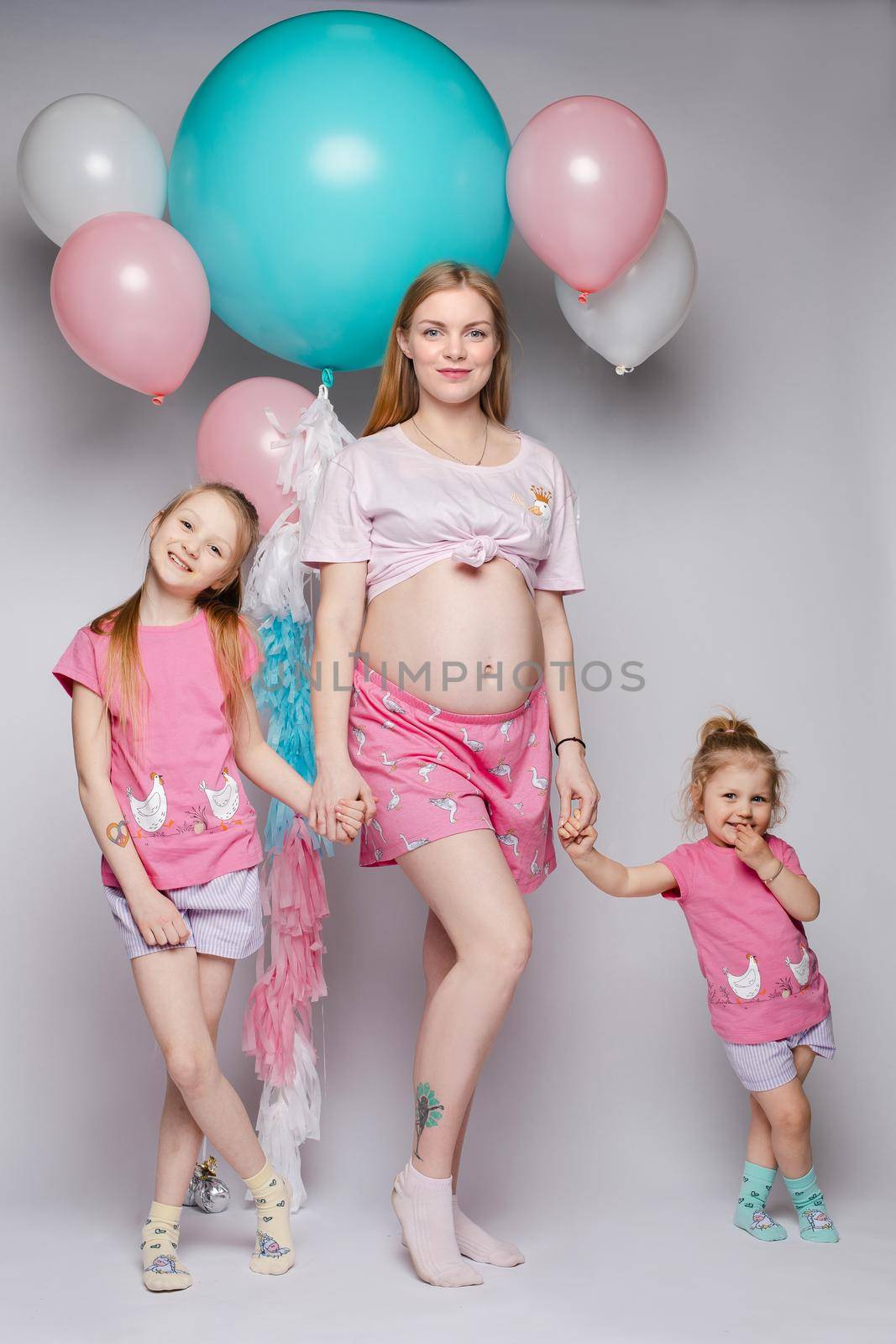 Stunning pregnant woman showing her belly. Her two daughters looking at their tummy too. They all wearing pyjamas. Studio portrait of mother and two daughters demonstrating bellies.