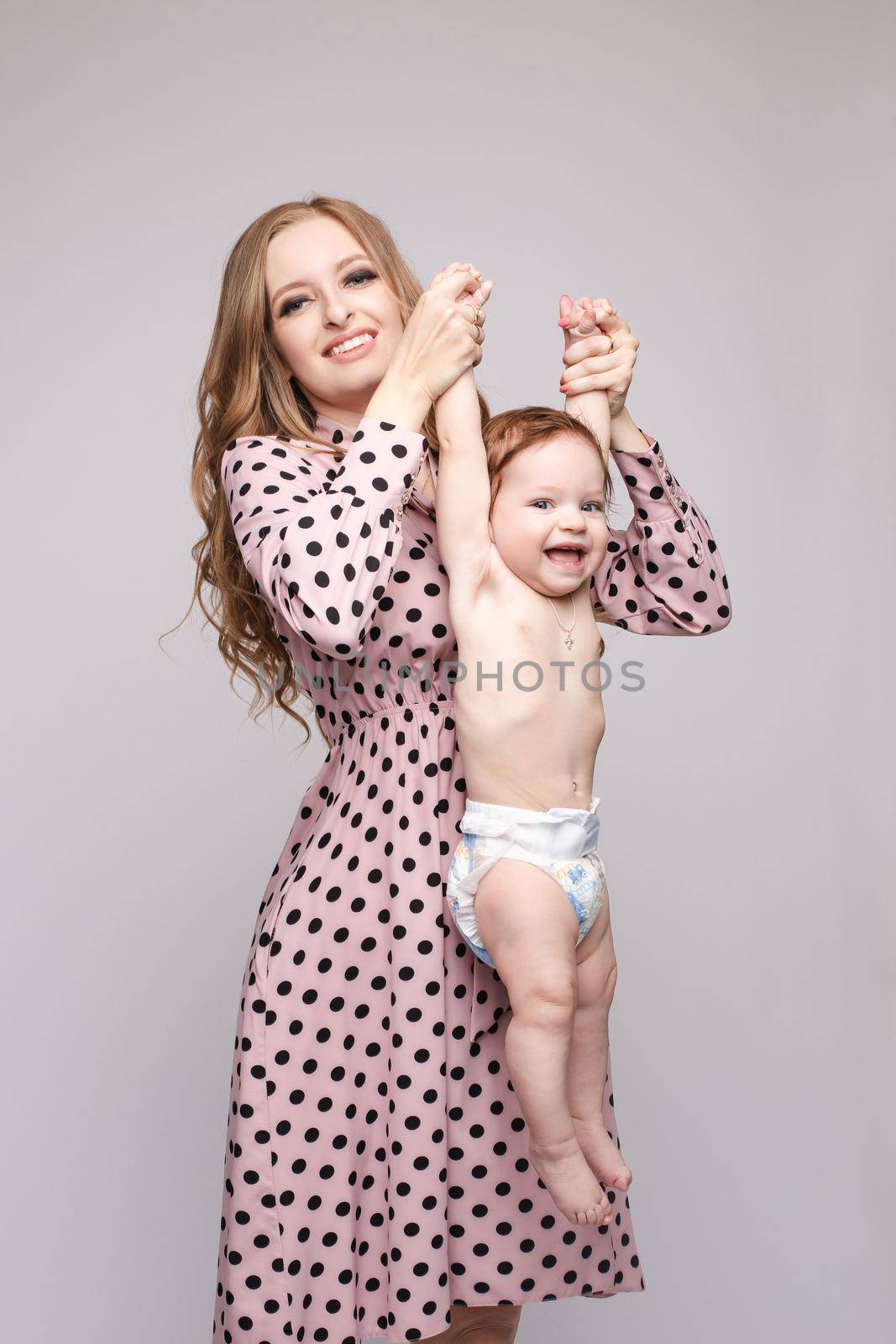 Front view of young mother keeping little child on hands and laughing on isolated background. Cheerful family posing and looking at camera in studio. Concept of happiness and childhood.