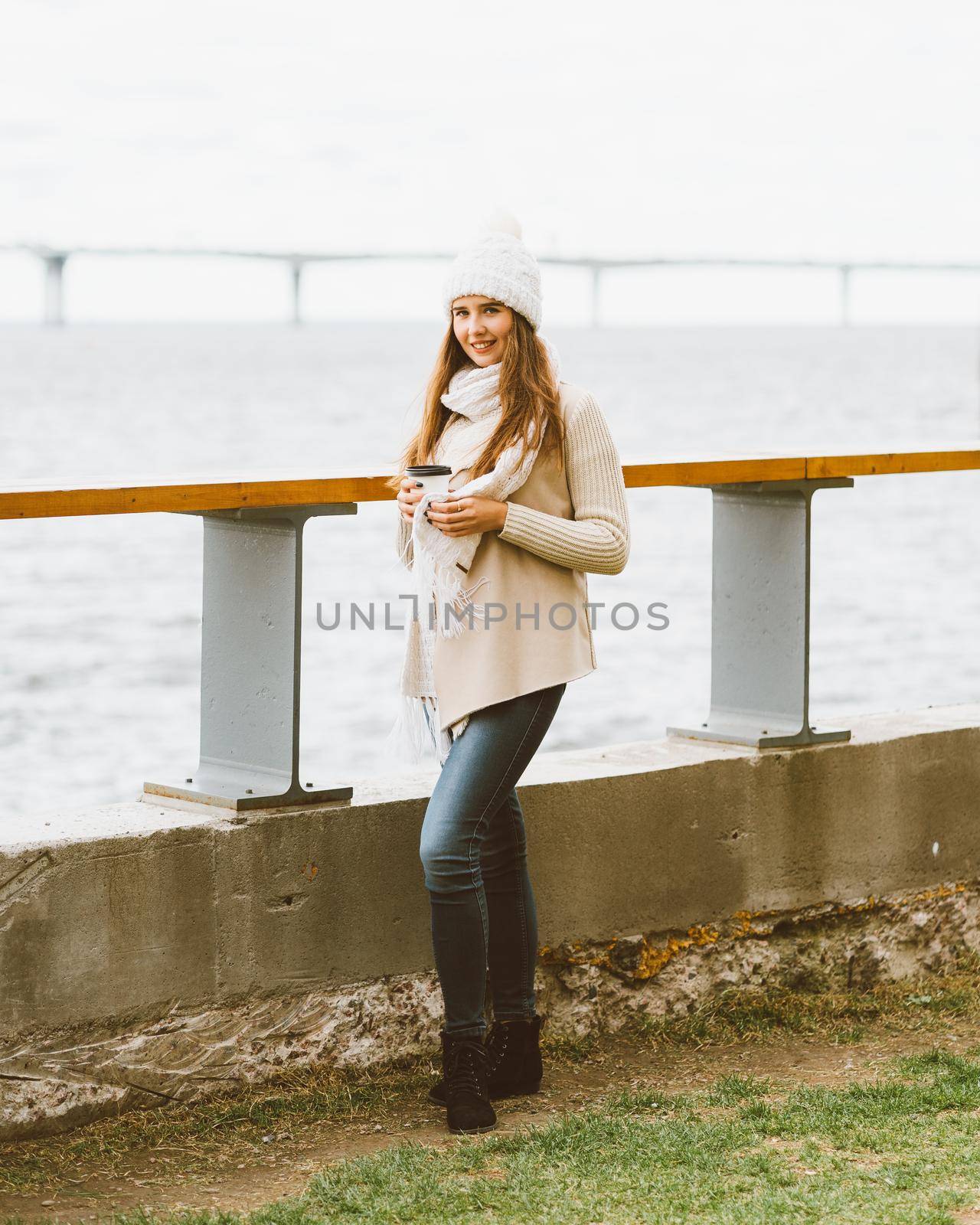 Beautiful young girl drinking coffee, tea from plastic mug in autumn, winter. A woman with long hair stands on waterfront on Baltic sea in port and waiting for ferry, heated by hot drink, copy space by NataBene