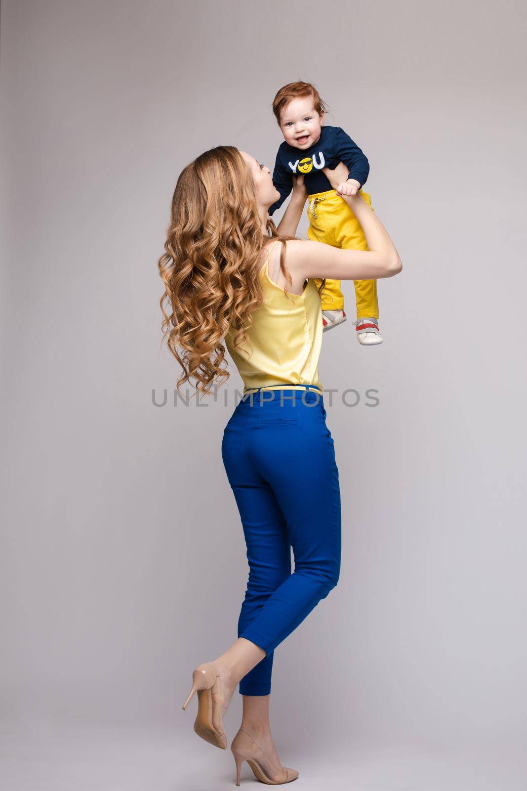 Young and fit woman in casual clothes holding little kid. Beautiful mother in yellow blouse and blue pants posing from back with her lovely son. Blonde mom on heels looking and happy smiling child.