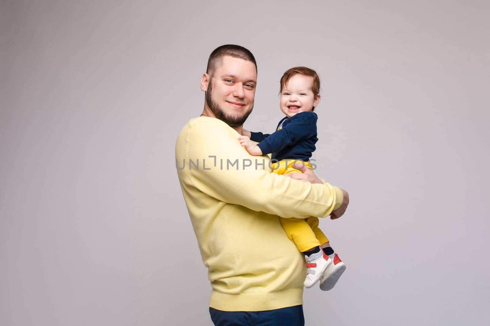 Portrait of happy father in yellow sweater posing with his lovely smiling child. Athletic and young men with beard holding red haired little boy. Handsome brunette dad hugging sweet and cute son.