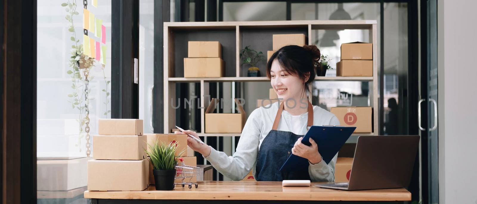 A portrait of a young Asian woman, e-commerce employee sitting in the office full of packages in the background write note of orders and a calculator, for SME business, e-commerce and delivery business.