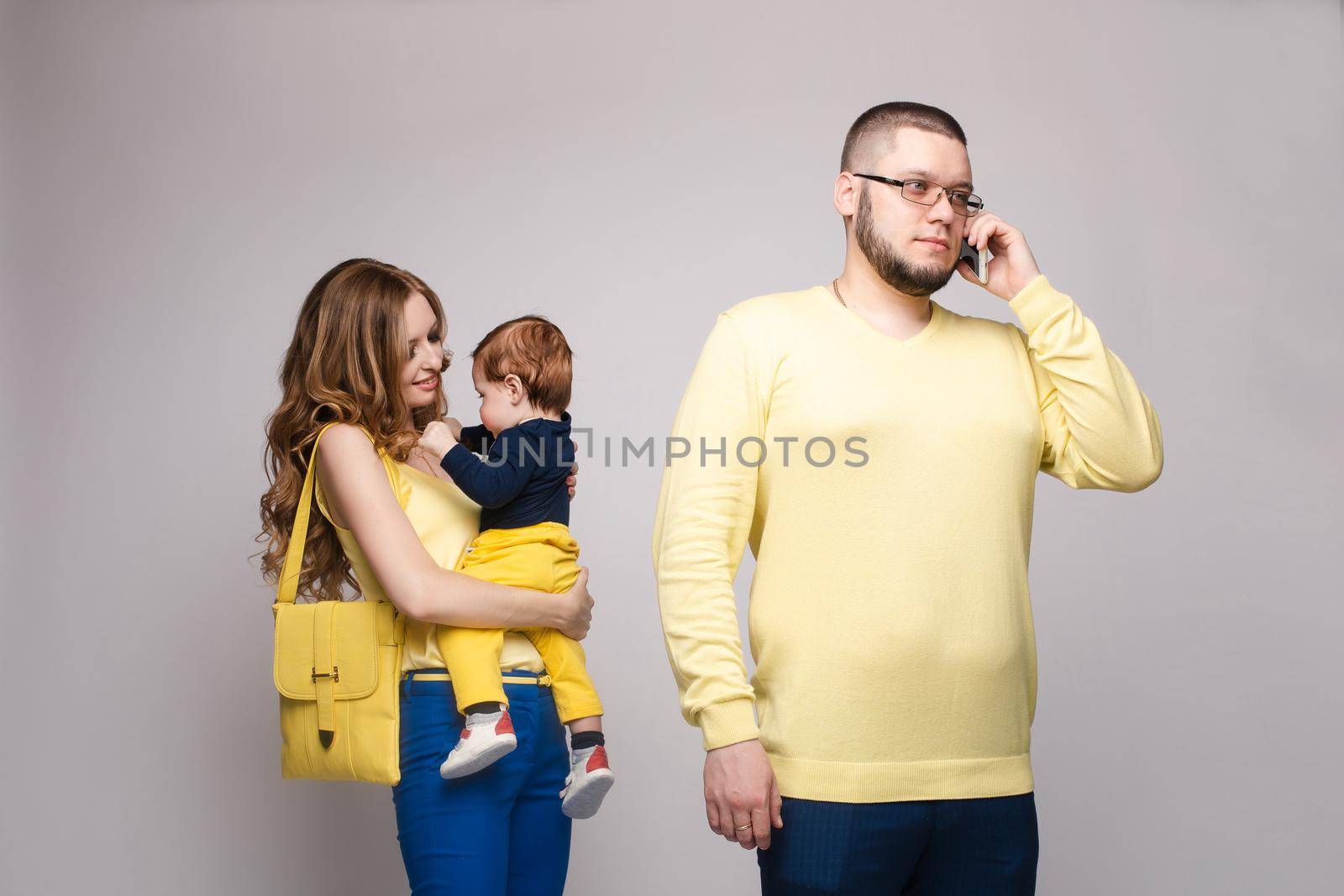 Busy father in glasses talking by phone while beautiful mother keeping little child on hands and playing behind. Pretty woman talking with kid and man working. Concept of parenthood.