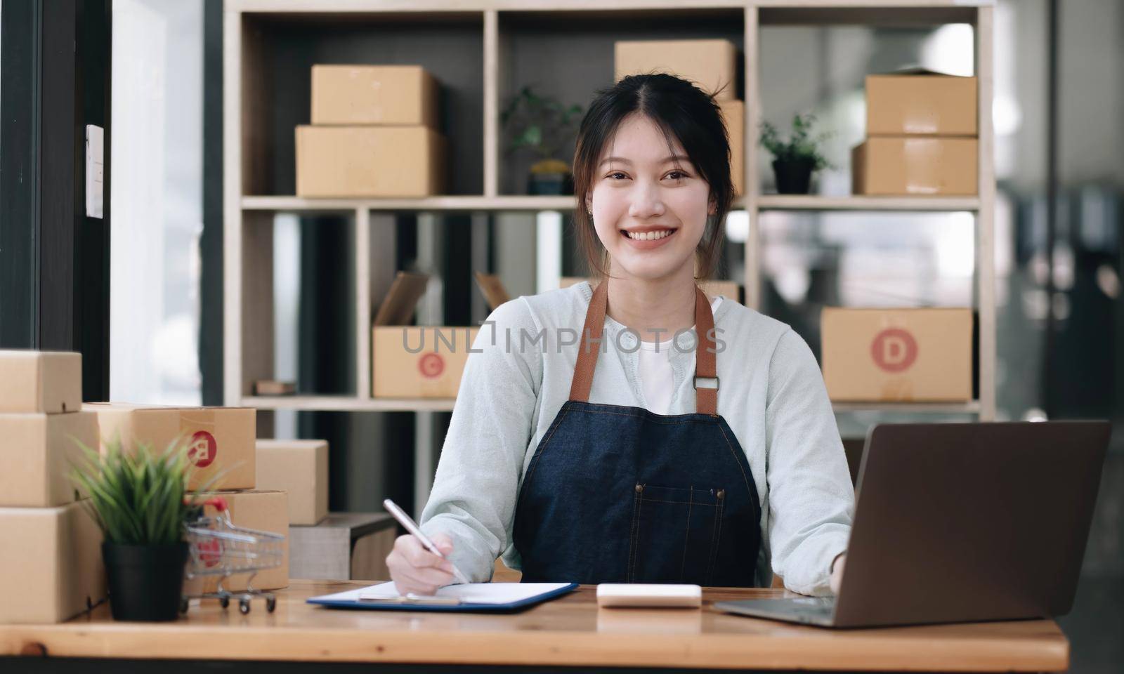 A portrait of a young Asian woman, e-commerce employee sitting in the office full of packages in the background write note of orders and a calculator, for SME business, e-commerce and delivery business. by wichayada