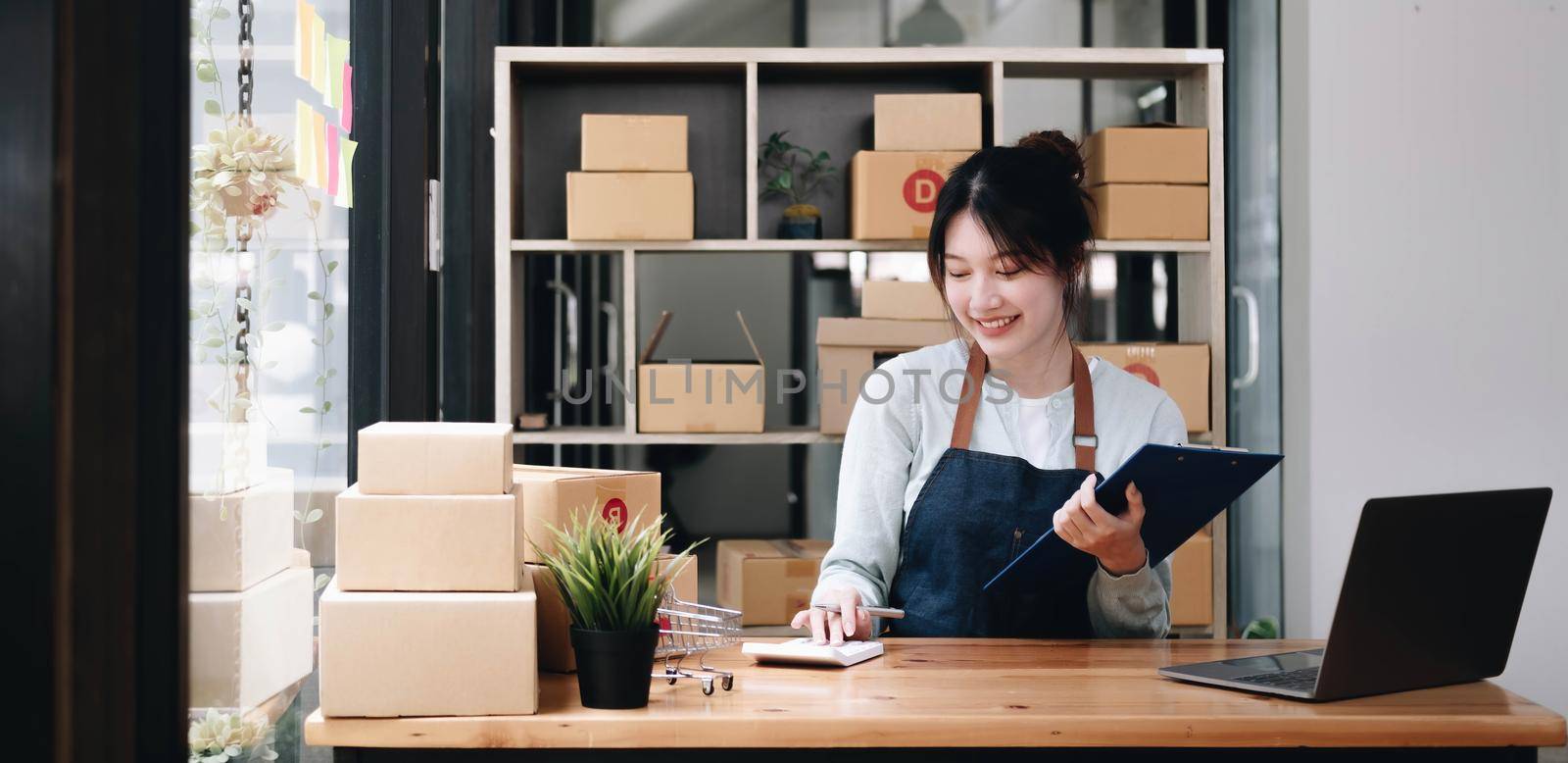 Smiling young Asian business owner woman prepare parcel box and standing check online orders of product for deliver to customer on laptop computer. Shopping Online concept..