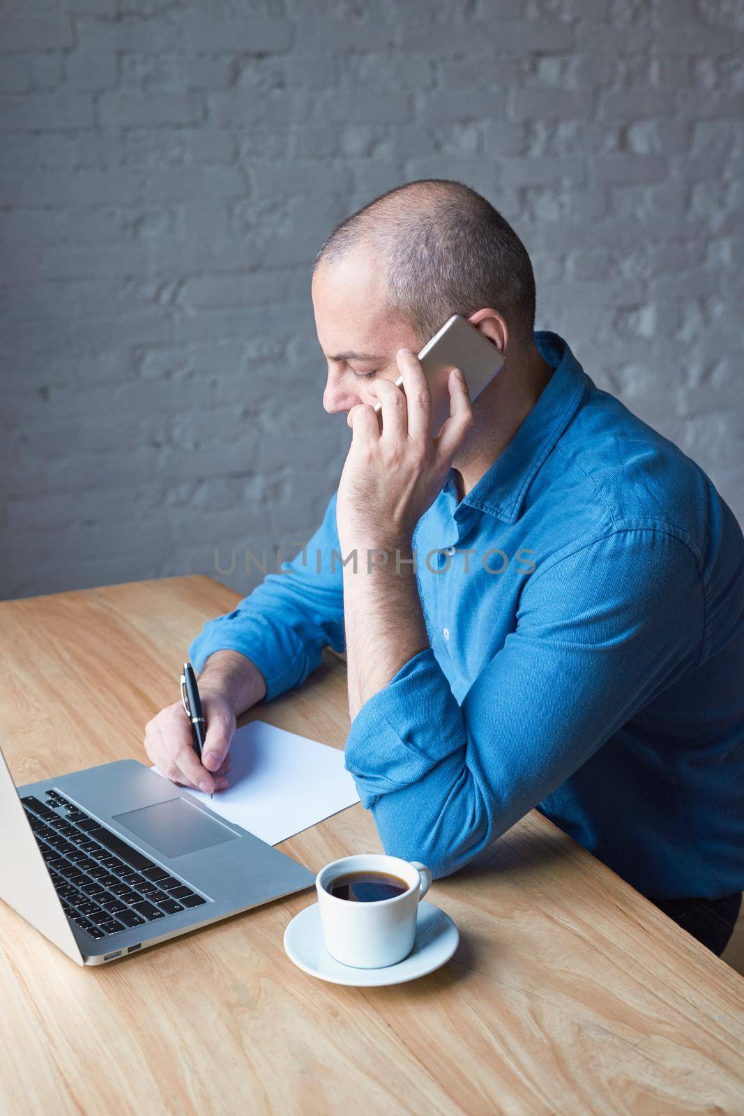 Handsome handsome mature man writes on a sheet of paper and talks on a cell phone, sits at a computer, laptop. Man with casual clothes in a blue shirt at a table in the office in front of the window by NataBene