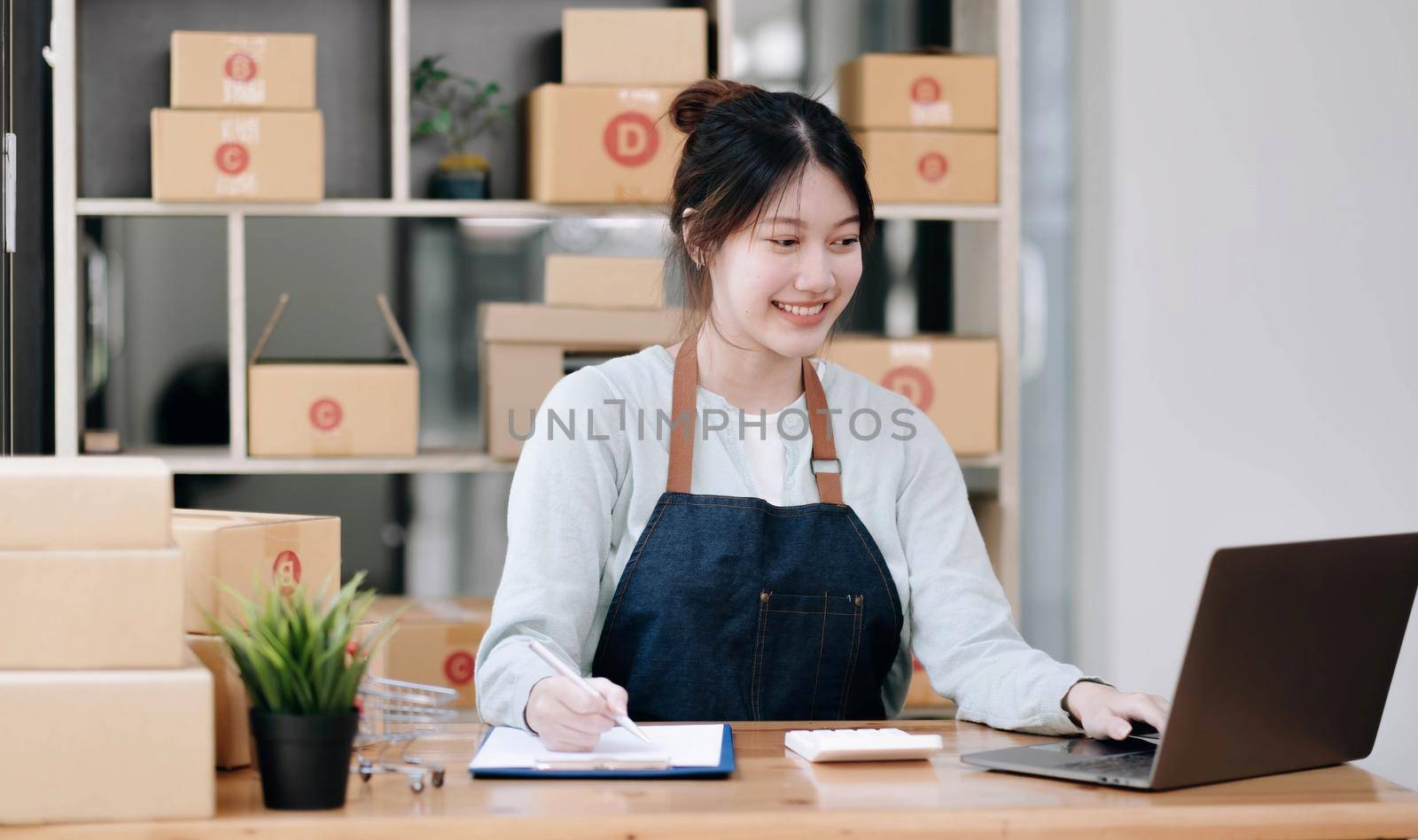 A portrait of a young Asian woman, e-commerce employee sitting in the office full of packages in the background write note of orders and a calculator, for SME business, e-commerce and delivery business. by wichayada