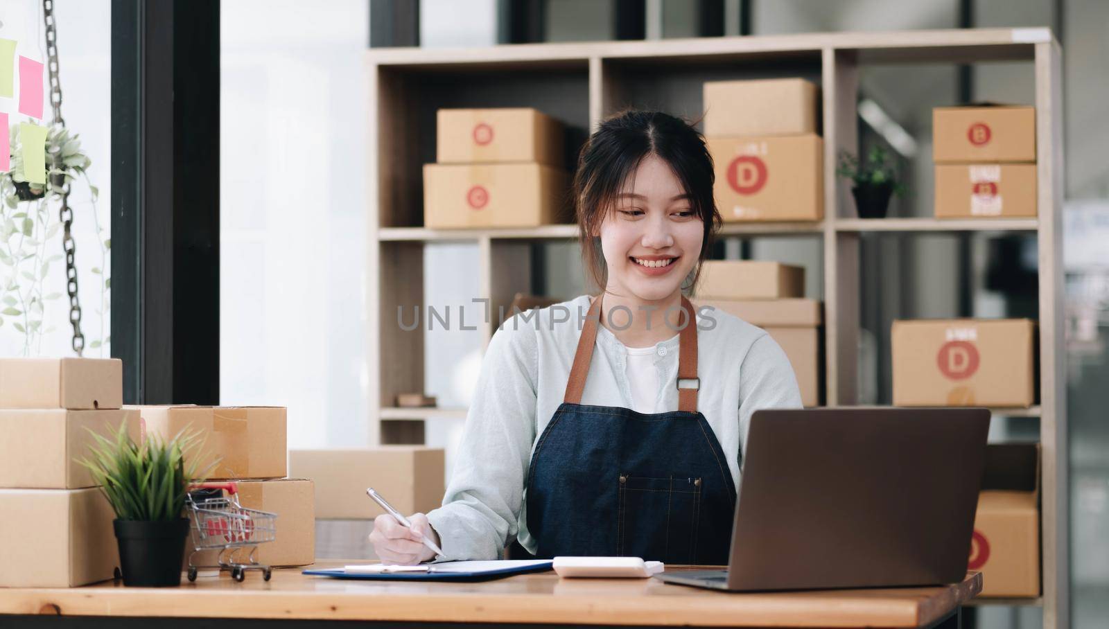 A portrait of a young Asian woman, e-commerce employee sitting in the office full of packages in the background write note of orders and a calculator, for SME business, e-commerce and delivery business. by wichayada