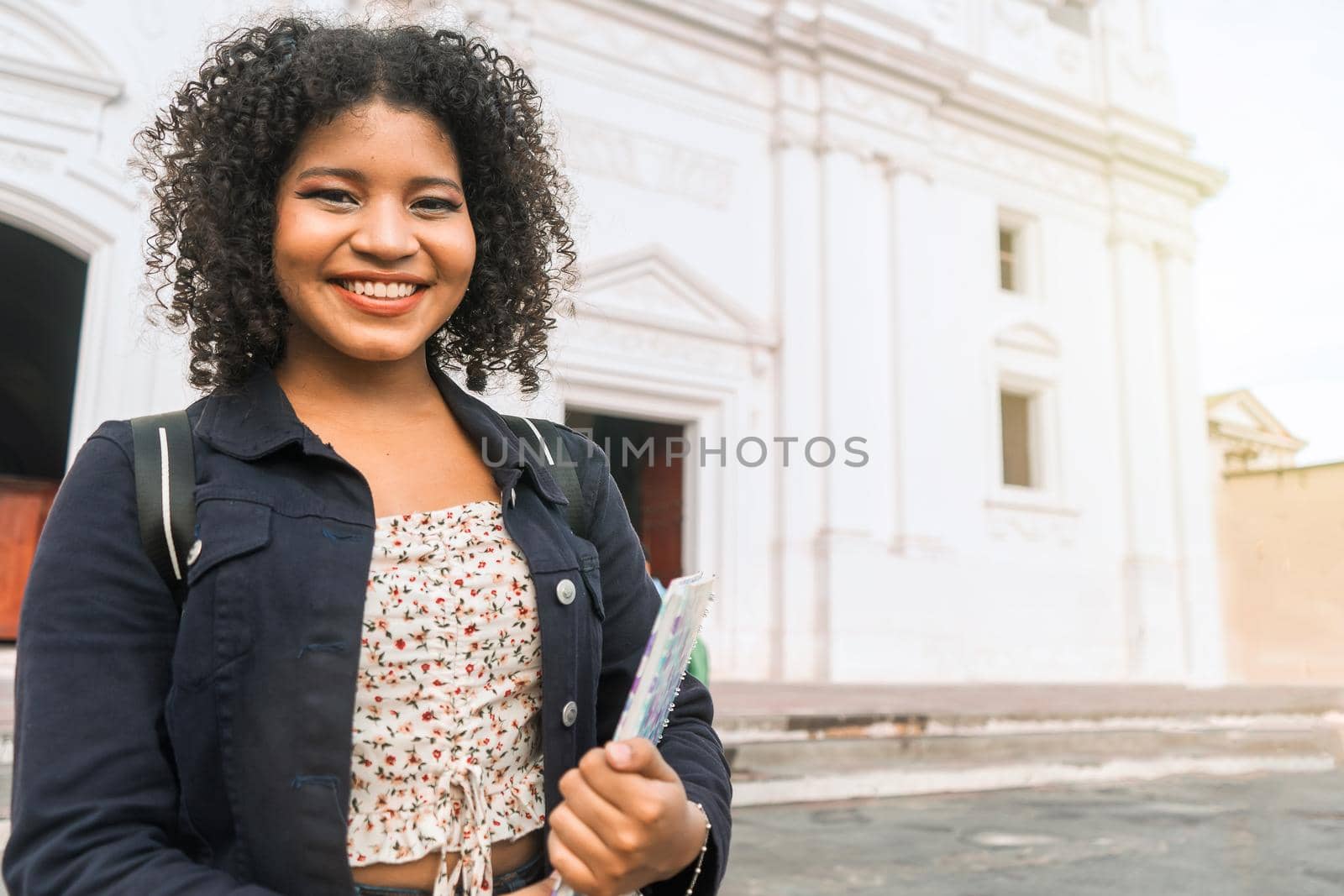 Curly-haired mestizo university student with a backpack and a notebook in her hand smiling and looking at the camera outdoors in the central park of leon nicaragua..