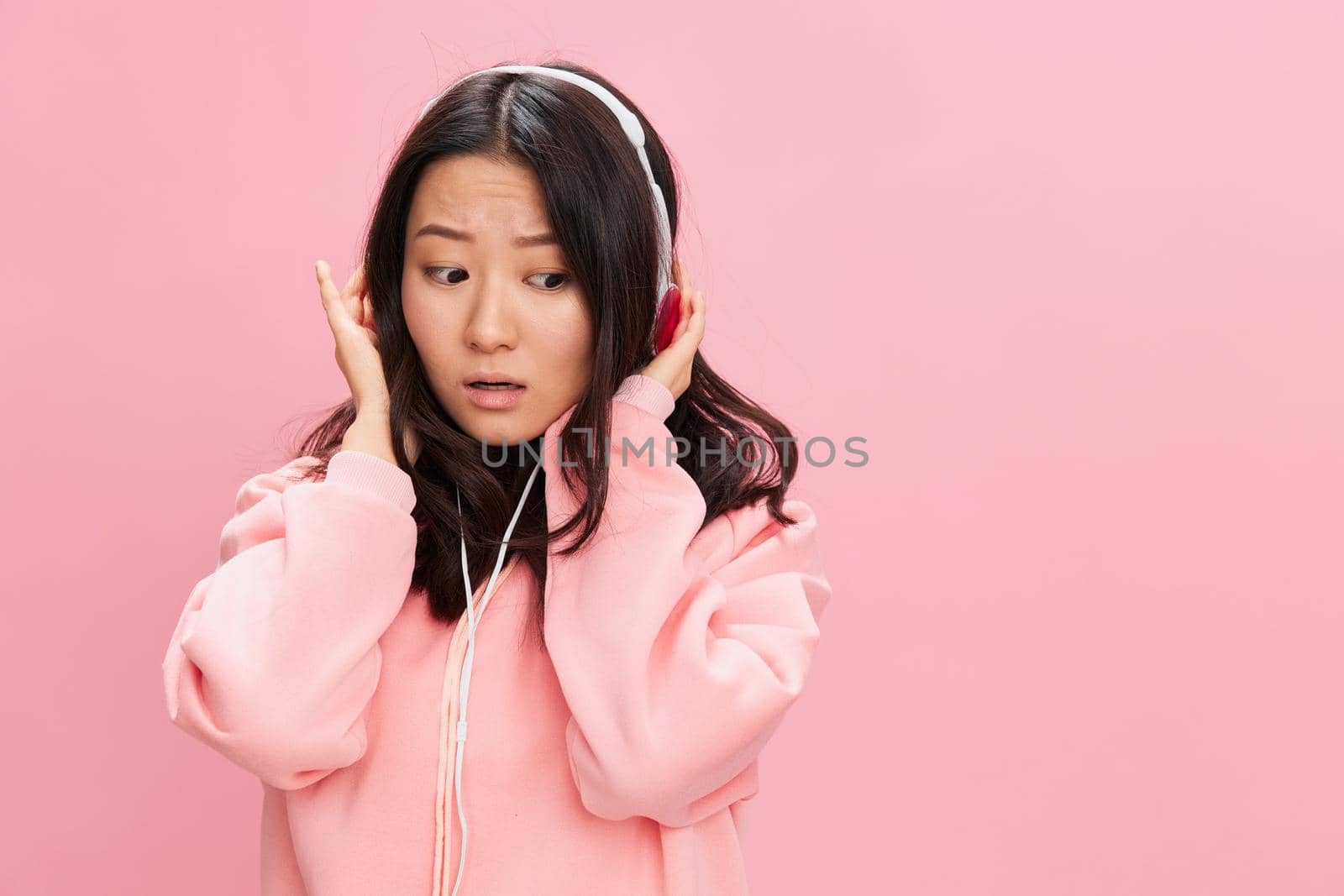 Upset confused Asian student young lady in pink hoodie sweatshirt with cute headphones looks at you posing isolated on over pink studio background. Good offer. Sound streaming platform ad concept