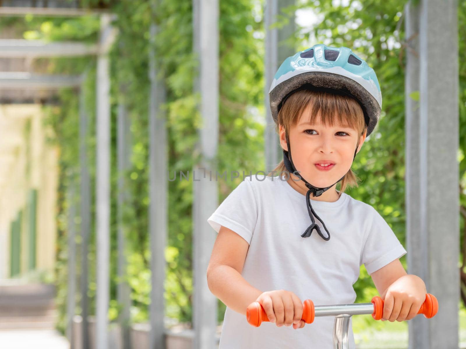 Little boy rides kick scooter in skate park. Portrait of kid in helmet on green leaves background. Training to skate at summer. by aksenovko