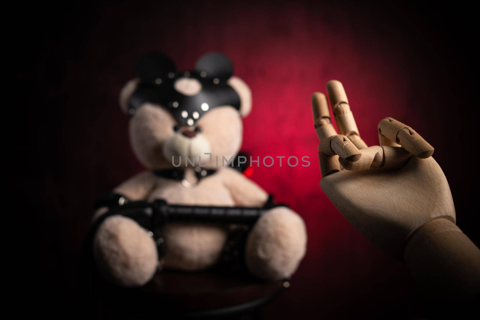 the toy teddy bear with a whip, dressed in leather belts and a mask, an accessory for BDSM games with a wooden hand and a sexual gesture
