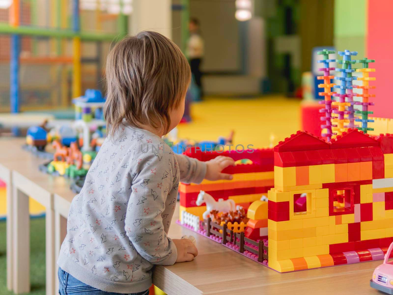 Toddler plays with colorful toy blocks. Little boy stares on figure from toy constructor. Interior of kindergarten or nursery. Indoors leisure activity for children.