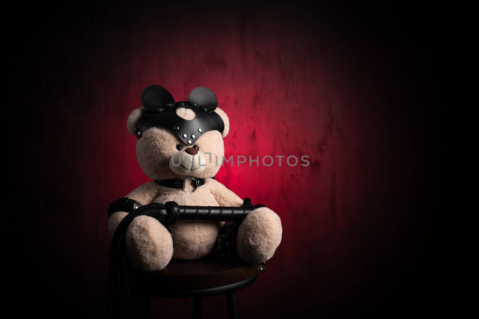 the toy teddy bear dressed in leather belts and a mask, an accessory for BDSM games on a dark red texture background
