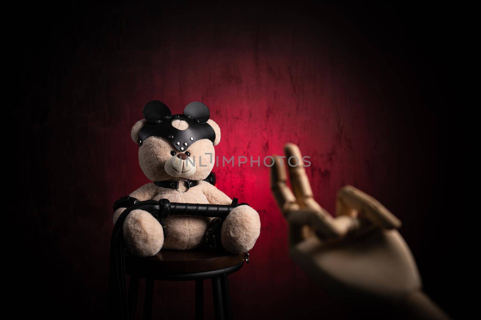 the toy teddy bear with a whip, dressed in leather belts and a mask, an accessory for BDSM games with a wooden hand and a sexual gesture