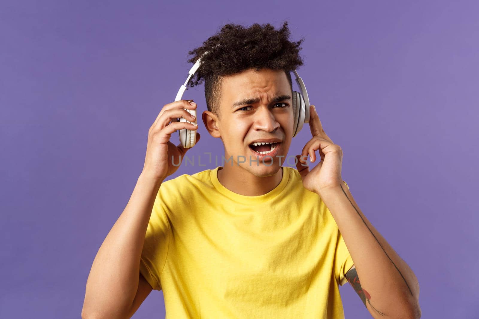 Cant hear you, repeat please. Portrait of young bothered guy interrupted of listening music, take-off headphones to answer person question, squinting look confused, purple background.