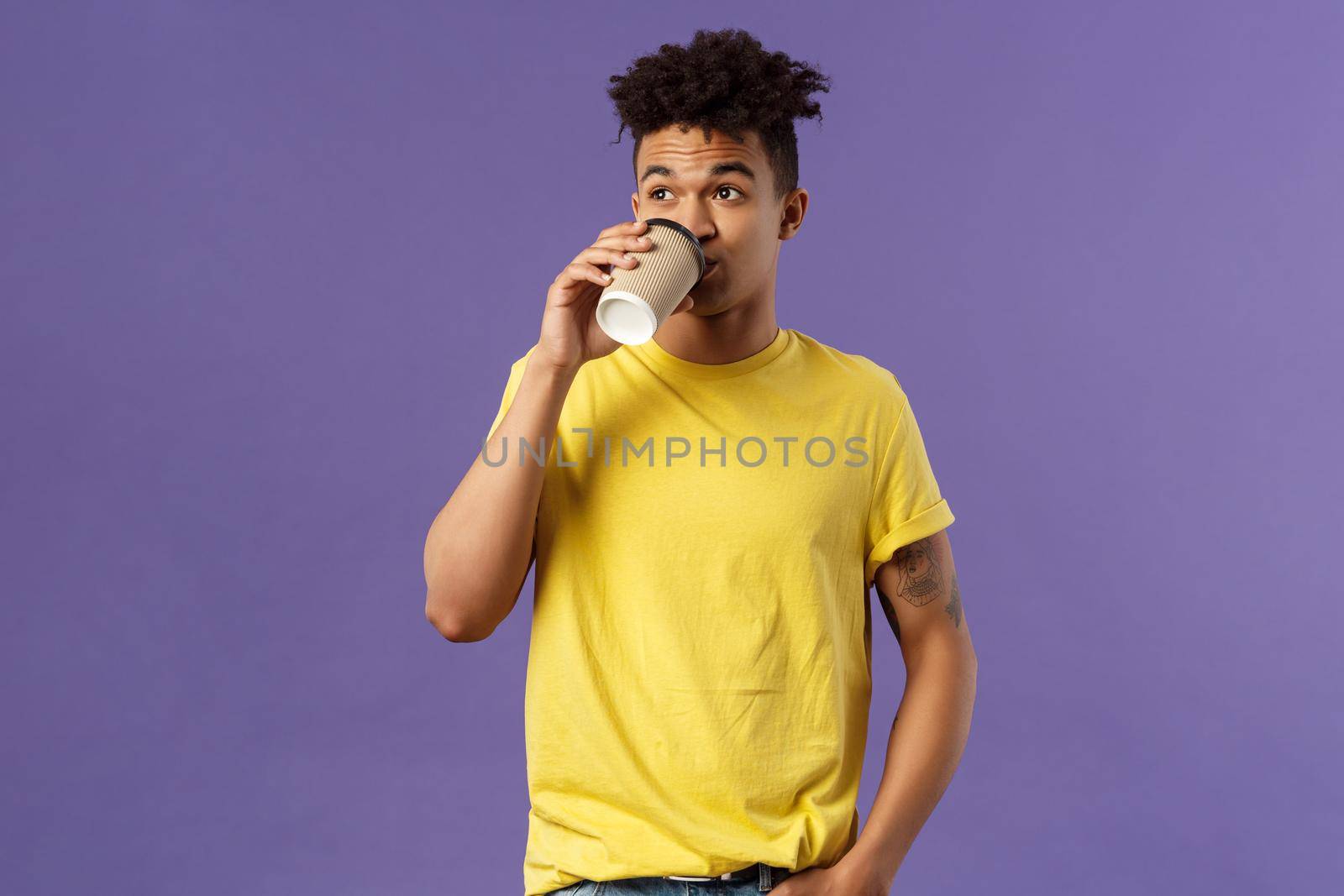 Portrait of carefree young male student getting energy in morning from caffeine, sipping coffee from take-away cup, look away casually standing, order drink in cafe, purple background.