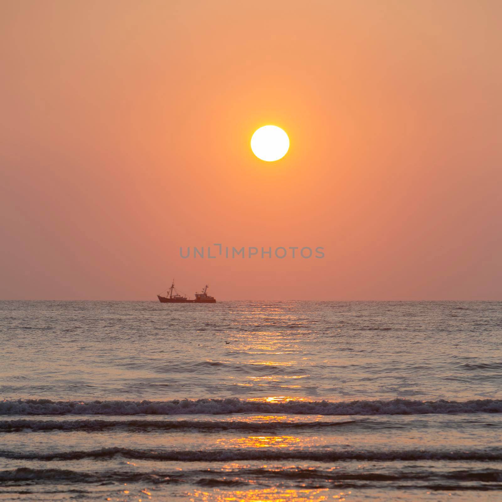 silhouette of fishing boat under orange sky and surf during sunset at sea seen from beach