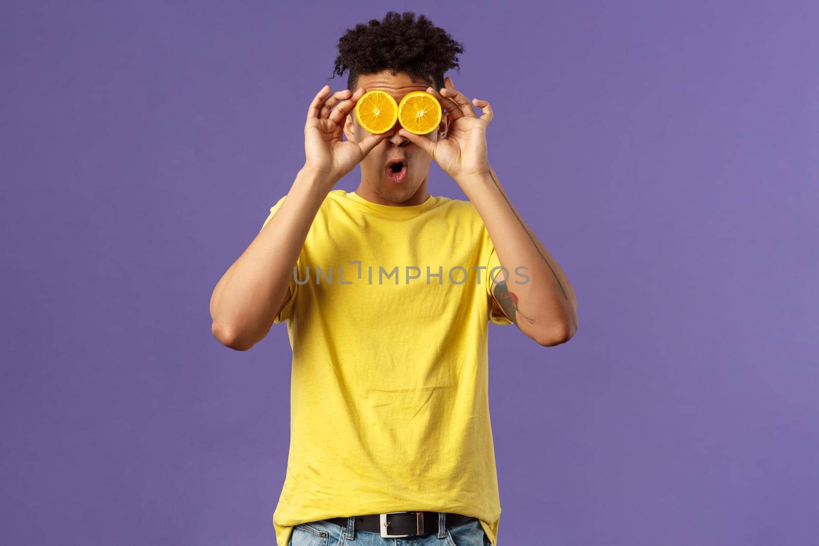 Holidays, vitamins and vacation concept. Portrait of funny, playful young guy fool around, playing with fruits, making eyes from pieces of orange, show wondered face expression, purple background.