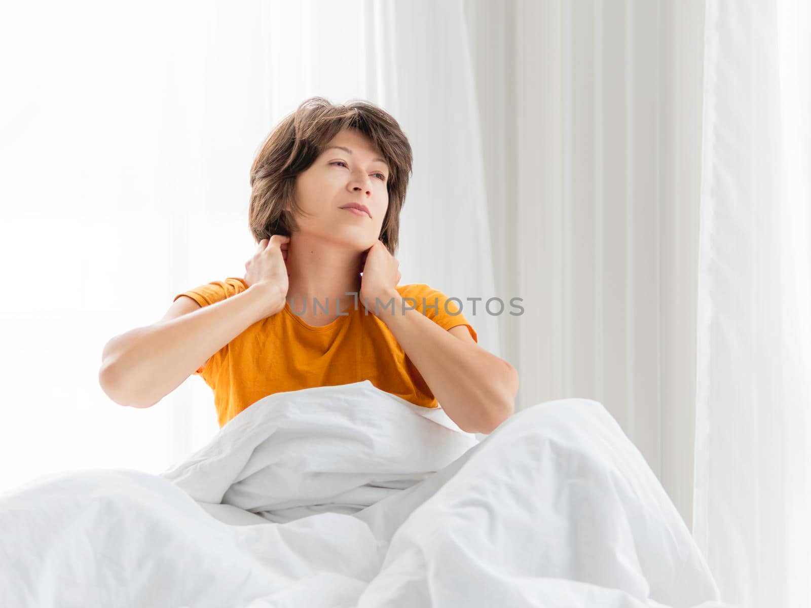 Sleepy woman in yellow pajama is massaging her neck. Waking up early in morning. Woman gets enough sleep. by aksenovko