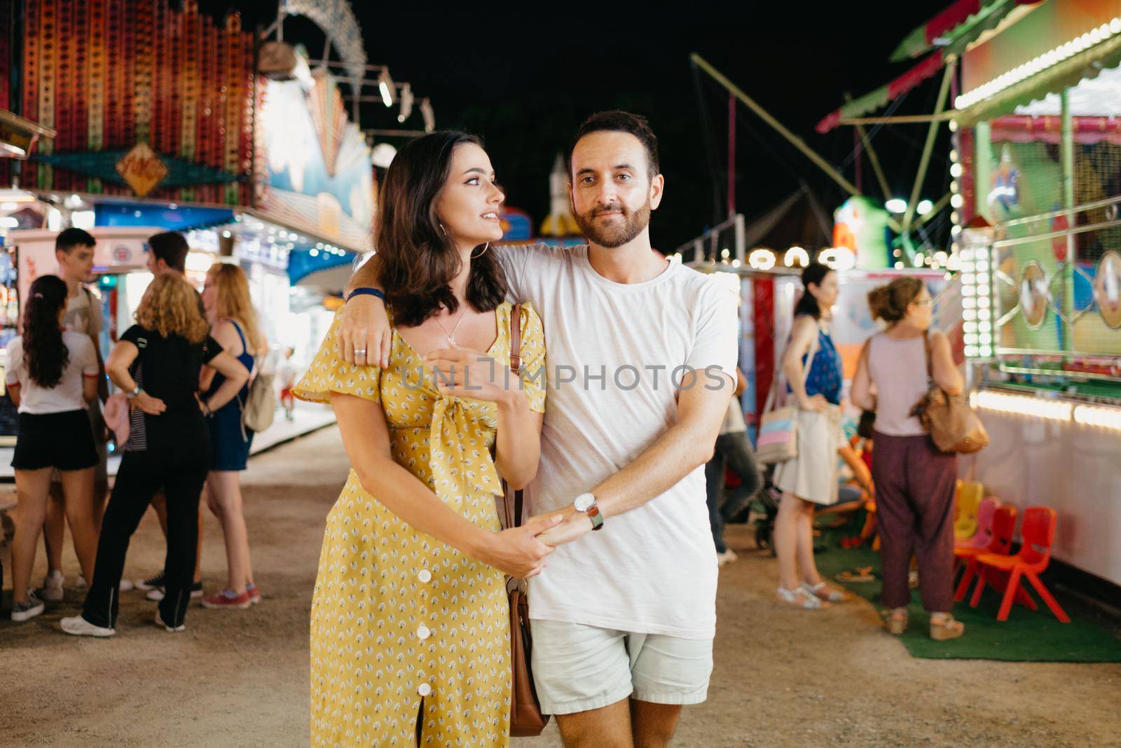 A woman in a yellow dress and her boyfriend with a beard are hugging each other and walking between amusement rides. A couple of lovers on a date at the fair in Valencia.