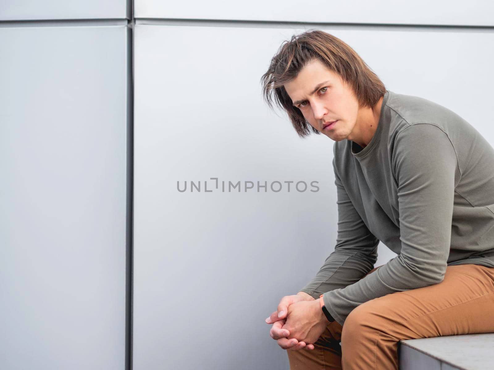 Millennial man with long hair and tense look sits near grey wall. Serious man in earth toned casual clothes is staring in camera. Copy space. by aksenovko