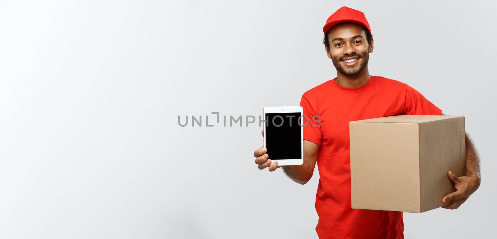 Delivery Concept - Portrait of Handsome African American delivery man or courier with box showing tablet on you to check the order. Isolated on Grey studio Background. Copy Space. by Benzoix