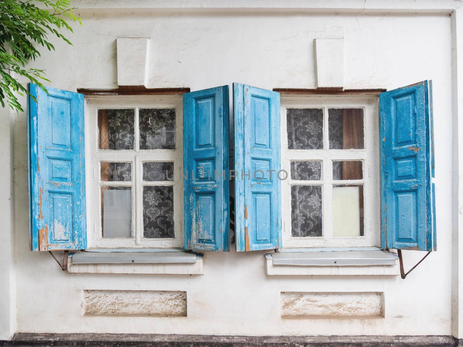 Old building with blue shutters on windows and old fashioned tulle curtains. White stone walls with retro architectural details. by aksenovko