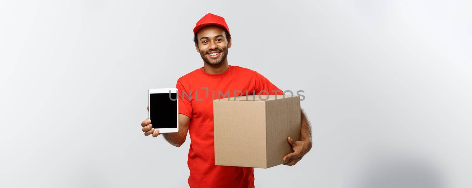 Delivery Concept - Portrait of Handsome African American delivery man or courier with box showing tablet on you to check the order. Isolated on Grey studio Background. Copy Space. by Benzoix