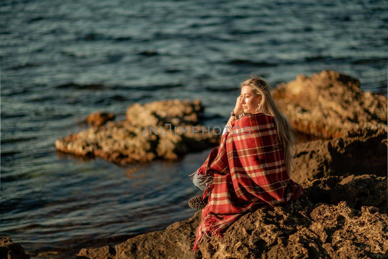 Attractive blonde Caucasian woman enjoying time on the beach at sunset, sitting in a blanket and looking to the side, with the sunset sky and sea in the background. Beach vacation