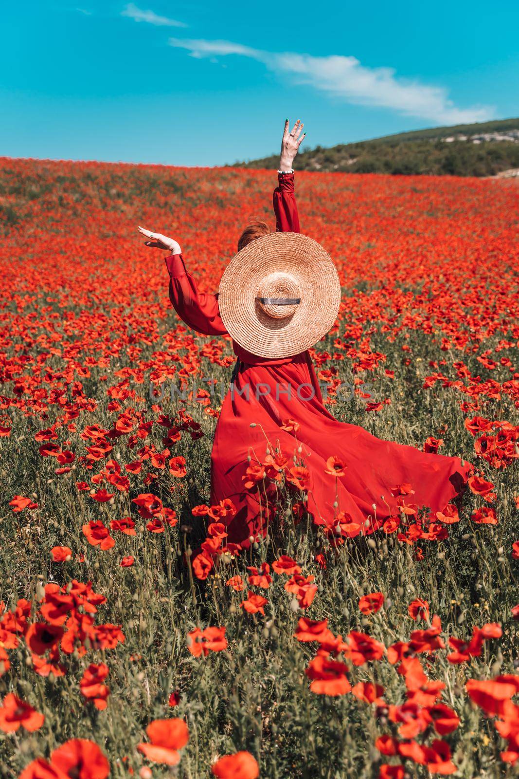 Young woman stands with her back in a long red dress and hat, posing on a large field of red poppies.