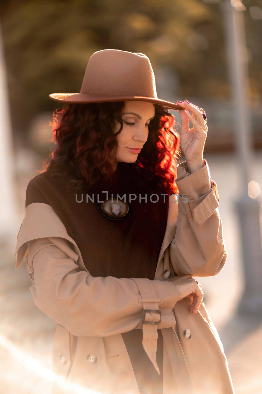 Outdoor fashion portrait of young elegant fashionable brunette woman, model in stylish hat, choker and light raincoat posing at sunset in European city