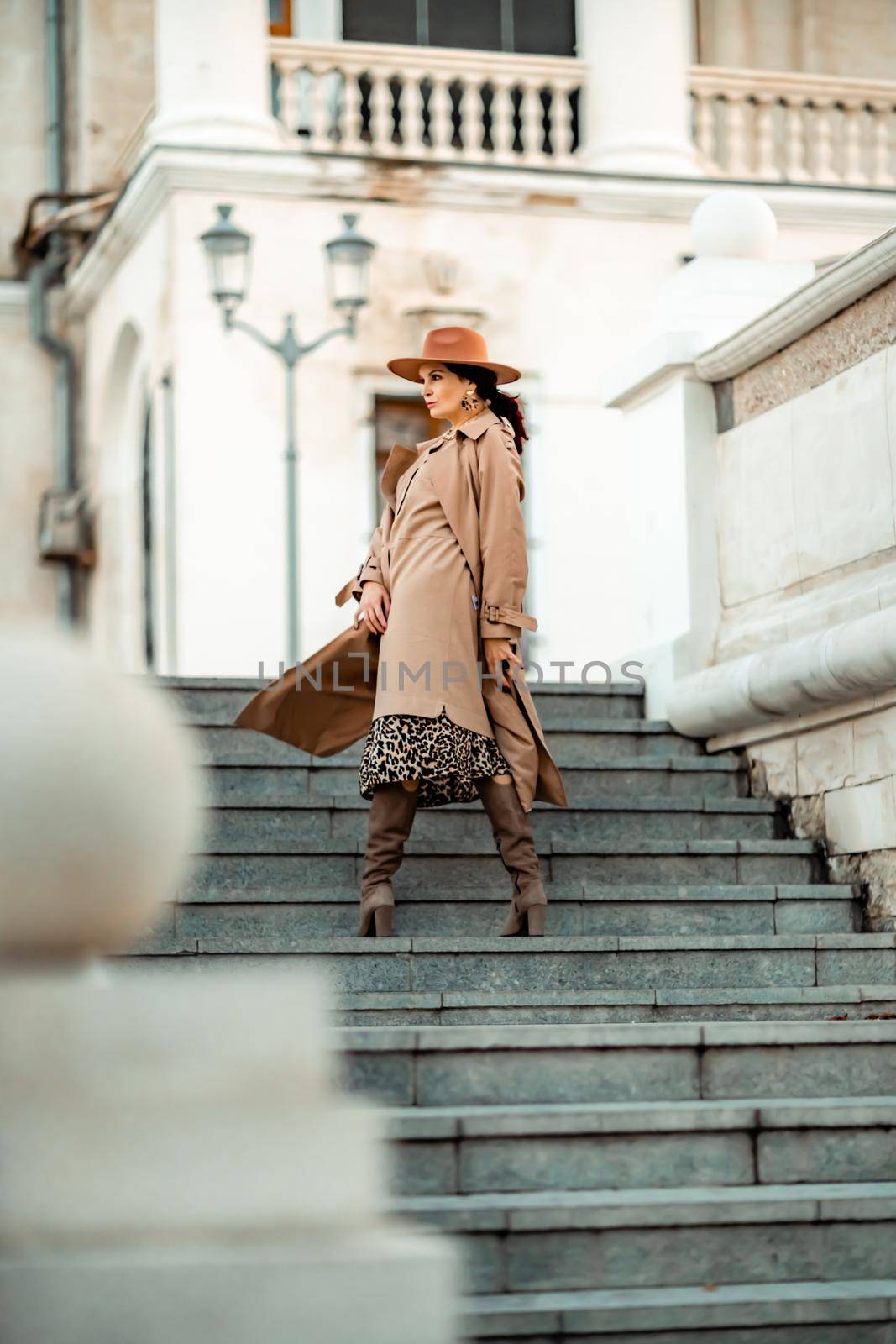 Outdoor fashion portrait of young elegant fashionable brunette woman, model in stylish hat, choker and light raincoat posing at sunset in European city. by Matiunina