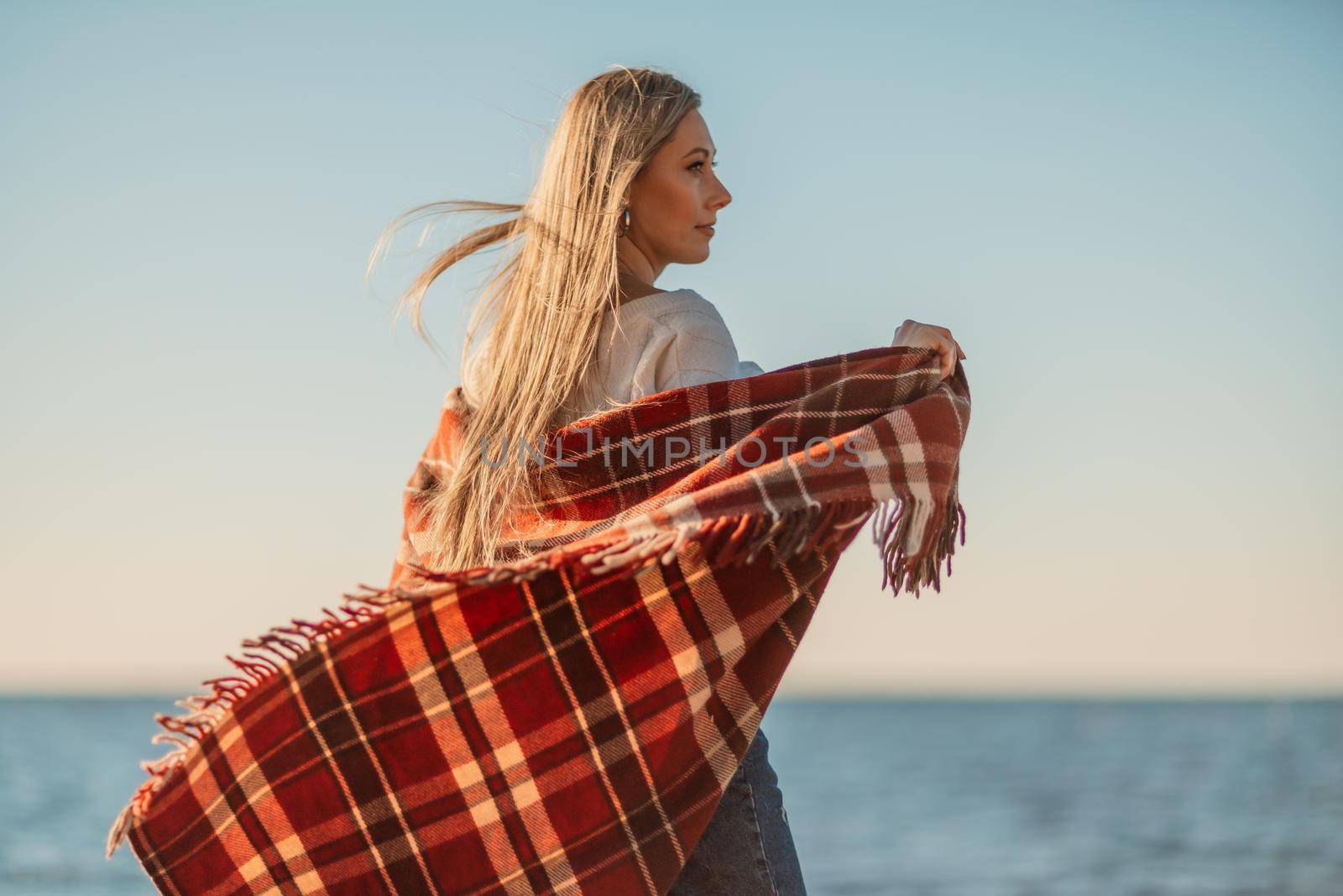 Attractive blonde Caucasian woman enjoying time on the beach at sunset, walking in a blanket and looking to the side, with the sunset sky and sea in the background. Beach vacation by Matiunina