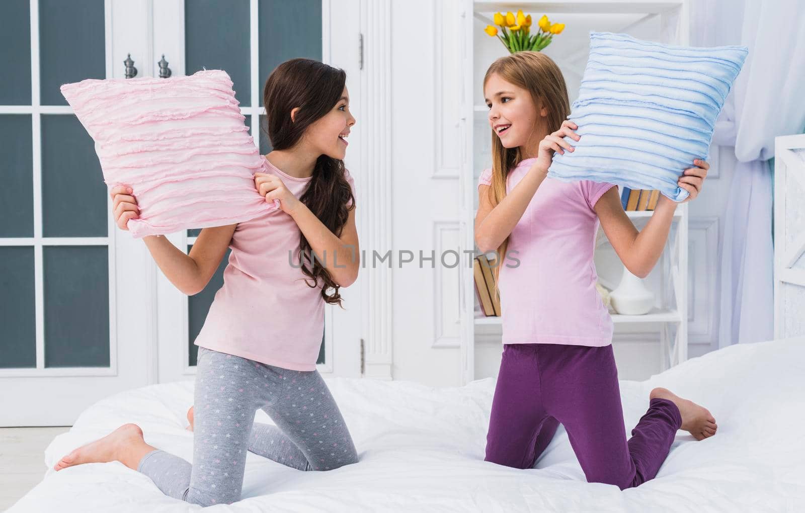 two happy female friends doing pillow fight bed home by Zahard