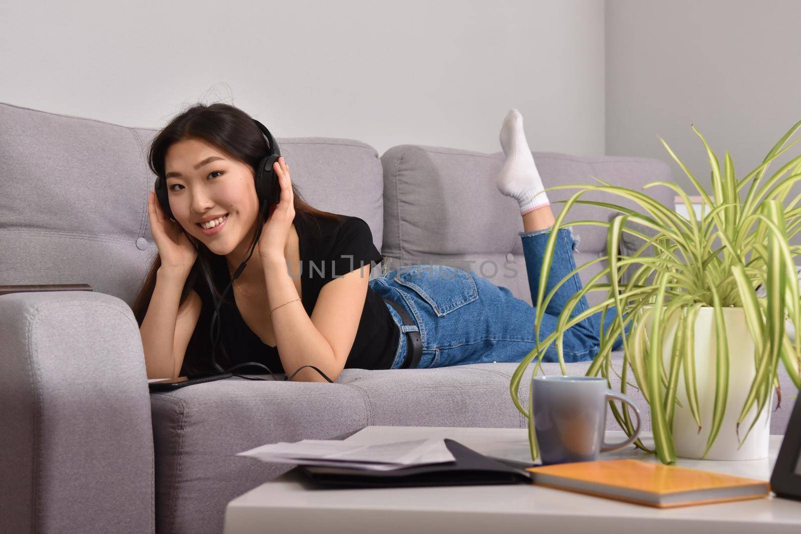 Excited beautiful asian teen listening music in headphones on her sofa at home. Wearing jeans and black tshirt