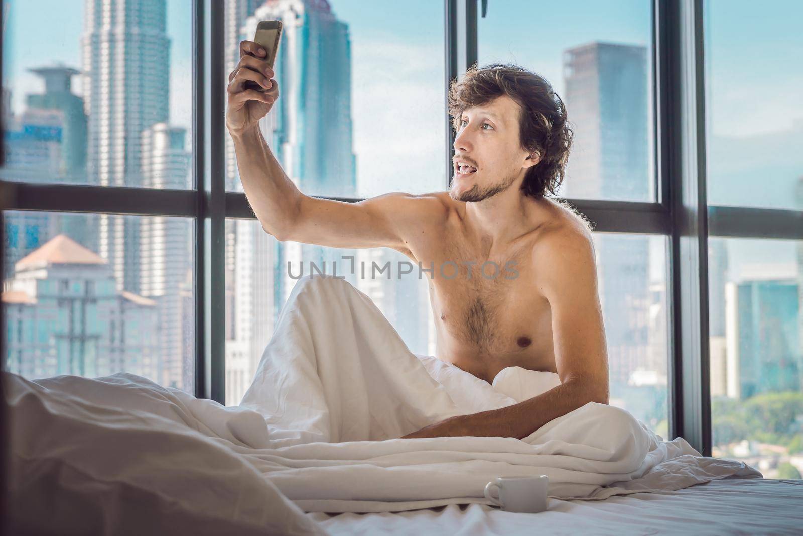 Man wakes up in the morning in an apartment in the downtown area with a view of the skyscrapers and uses smartphone. Life in the noise of the big city concept. Not enough sleep.