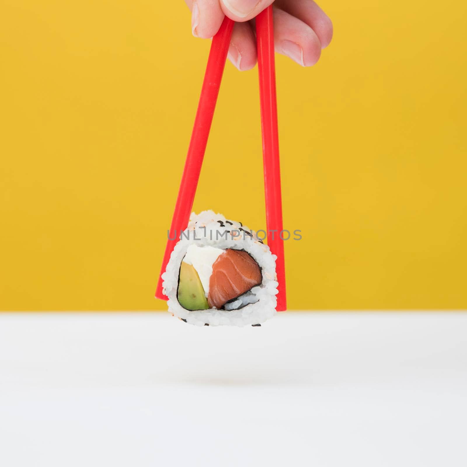 close up person s hand holding sushi with red chopsticks against yellow backdrop