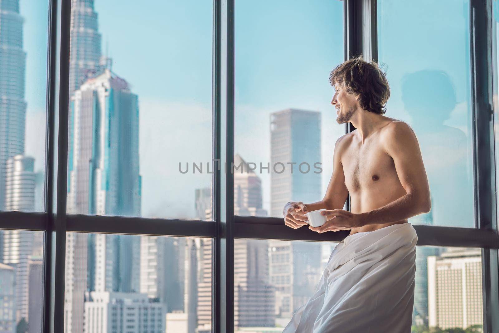 Man wakes up in the morning in an apartment in the downtown area with a view of the skyscrapers and drinks coffee. Life in the noise of the big city concept. Not enough sleep by galitskaya